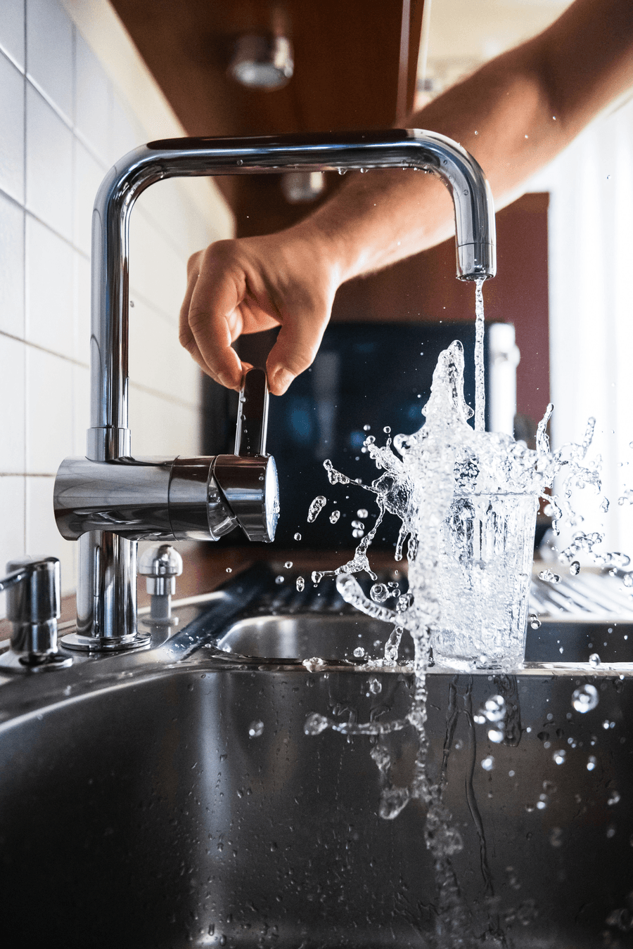Mão abrindo a torneira da pia, com água enchendo um copo sobre a bancada de inox — serviços hidráulicos da JC Pequenos Reparos
